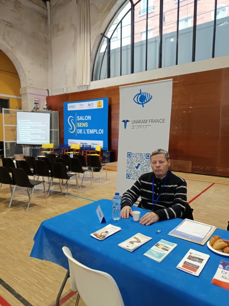 Joël Betton, à la table du stand et devant le kakémono de l'UNAKAM avant l'arrivée massive des visiteurs. Sur sa gauche : l'espace des conférences du Salon.